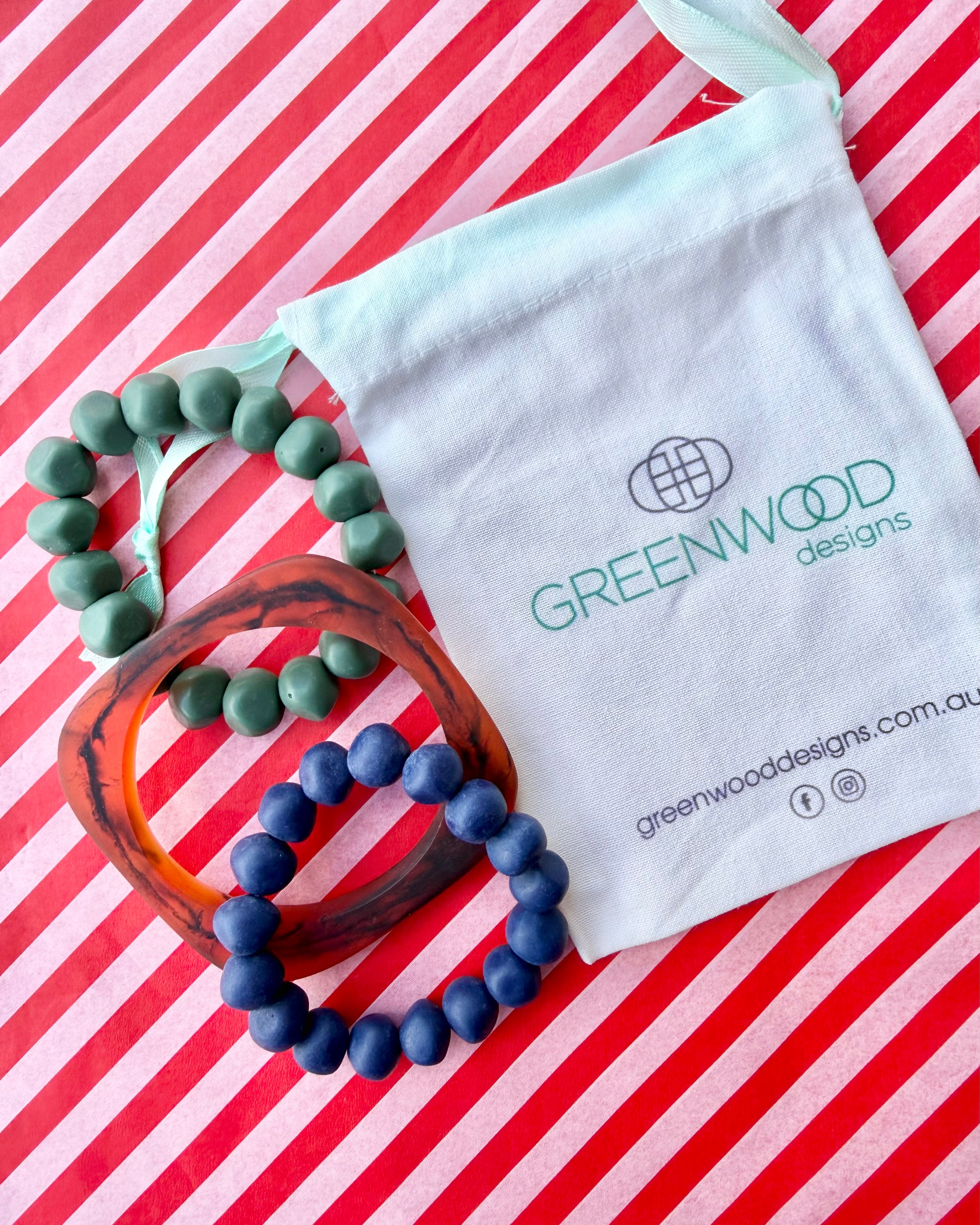 A muted green rock bracelet, a geo tortoiseshell bangle and a navy blue rock bracelet laid on red and white striped paper with a Greenwood Designs calico gift bag.
