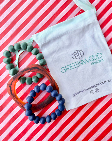 A muted green rock bracelet, a geo tortoiseshell bangle and a navy blue rock bracelet laid on red and white striped paper with a Greenwood Designs calico gift bag.