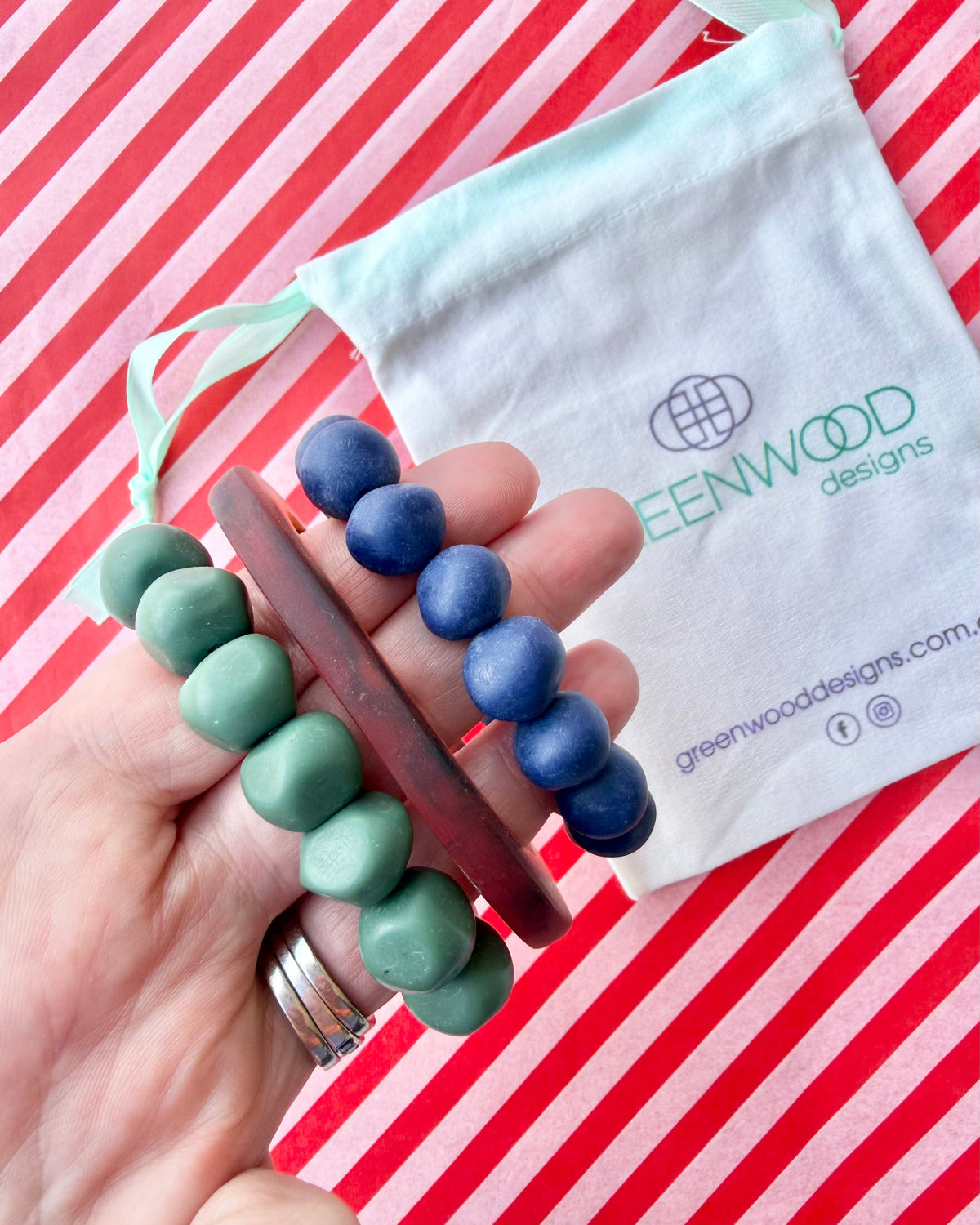 Hand holding a muted green rock bracelet, a slim tortoiseshell bangle and a navy blue rock bracelet, with a Greenwood Designs calico gift bag.