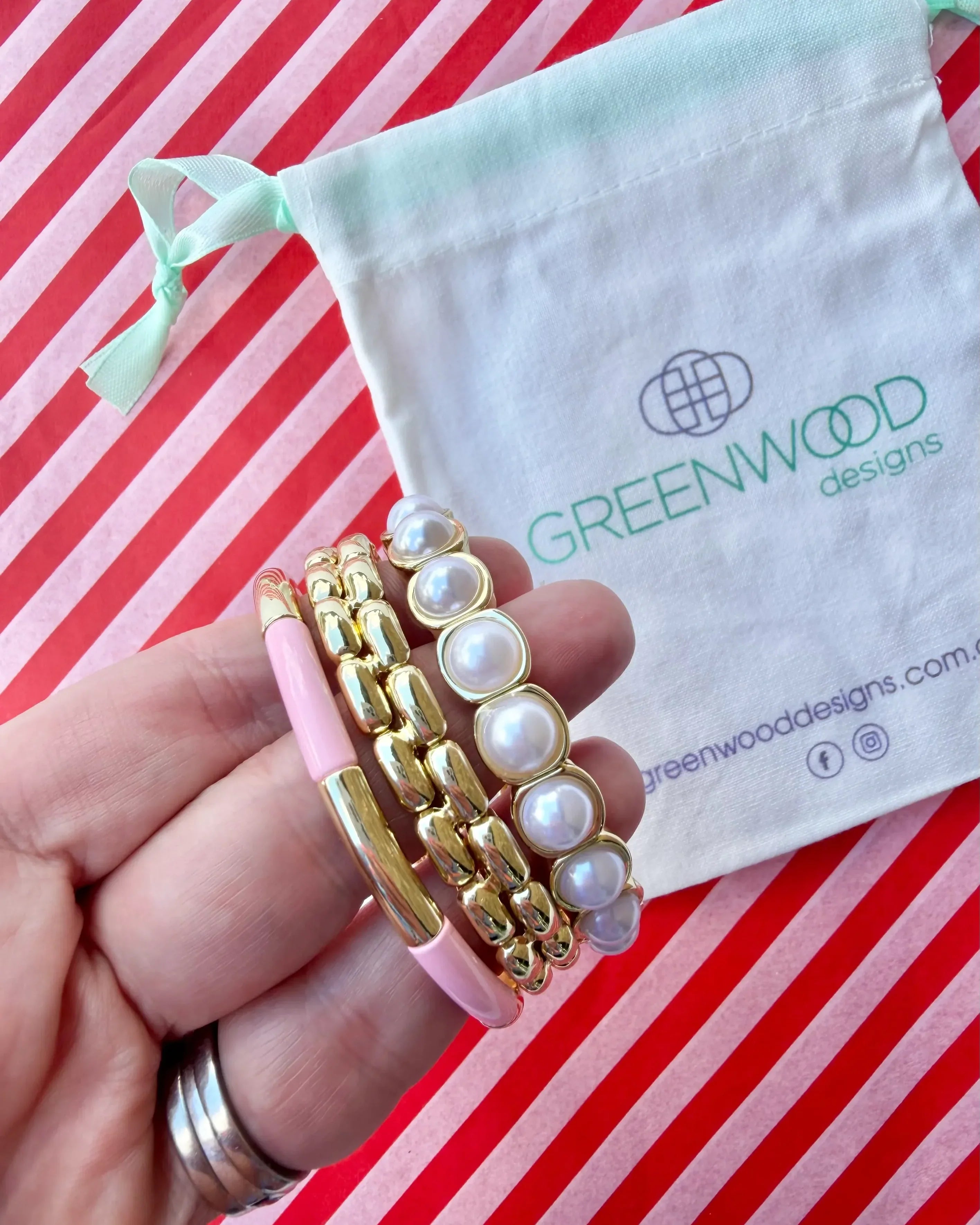 Hand holding three stacked bracelets: a pale pink enamel tube bracelet with a gold bar, a chunky gold link bracelet and a gold-framed pearl bracelet, with a Greenwood Designs calico gift bag.