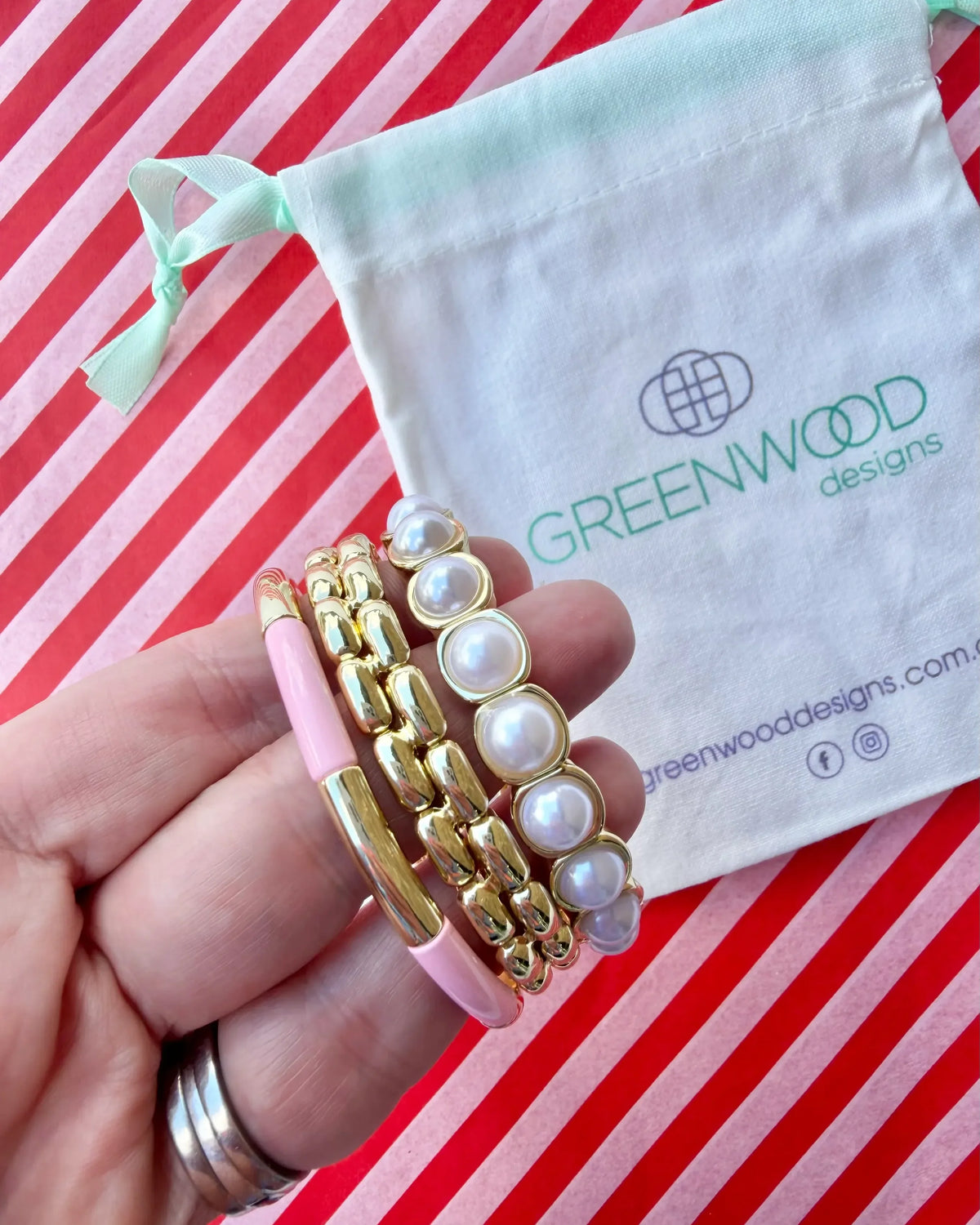 Hand holding three stacked bracelets: a pale pink enamel tube bracelet with a gold bar, a chunky gold link bracelet and a gold-framed pearl bracelet, with a Greenwood Designs calico gift bag.