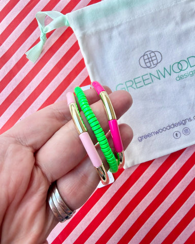 Hand holding three stacked bracelets: a pale pink enamel tube bracelet with a gold bar, a neon green beaded bracelet and a hot pink enamel tube bracelet with a gold bar, with a Greenwood Designs calico gift bag.