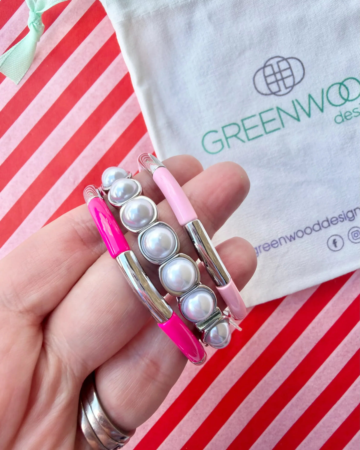 Hand holding three stacked bracelets: a hot pink enamel tube bracelet, a silver-framed pearl bracelet and a pale pink enamel tube bracelet, with a Greenwood Designs calico gift bag.