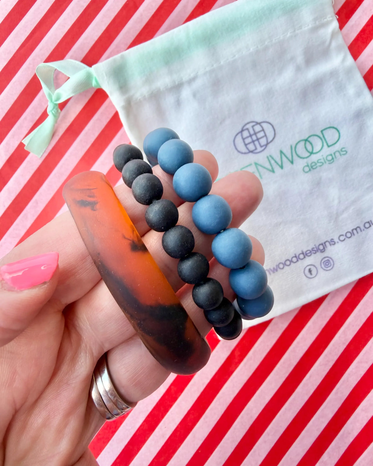 Hand holding a chunky tortoiseshell bangle, a matte black bead bracelet and a dusty blue bead bracelet, with a Greenwood Designs calico gift bag.