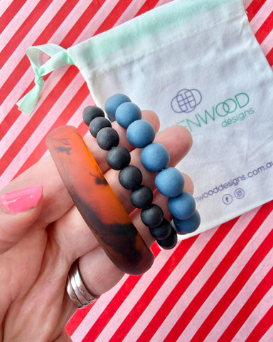 Hand holding a chunky tortoiseshell bangle, a matte black bead bracelet and a dusty blue bead bracelet, with a Greenwood Designs calico gift bag.