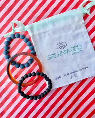 A dusty blue bead bracelet, a tortoiseshell bangle and a matte black bead bracelet laid on red and white striped paper with a Greenwood Designs calico gift bag.