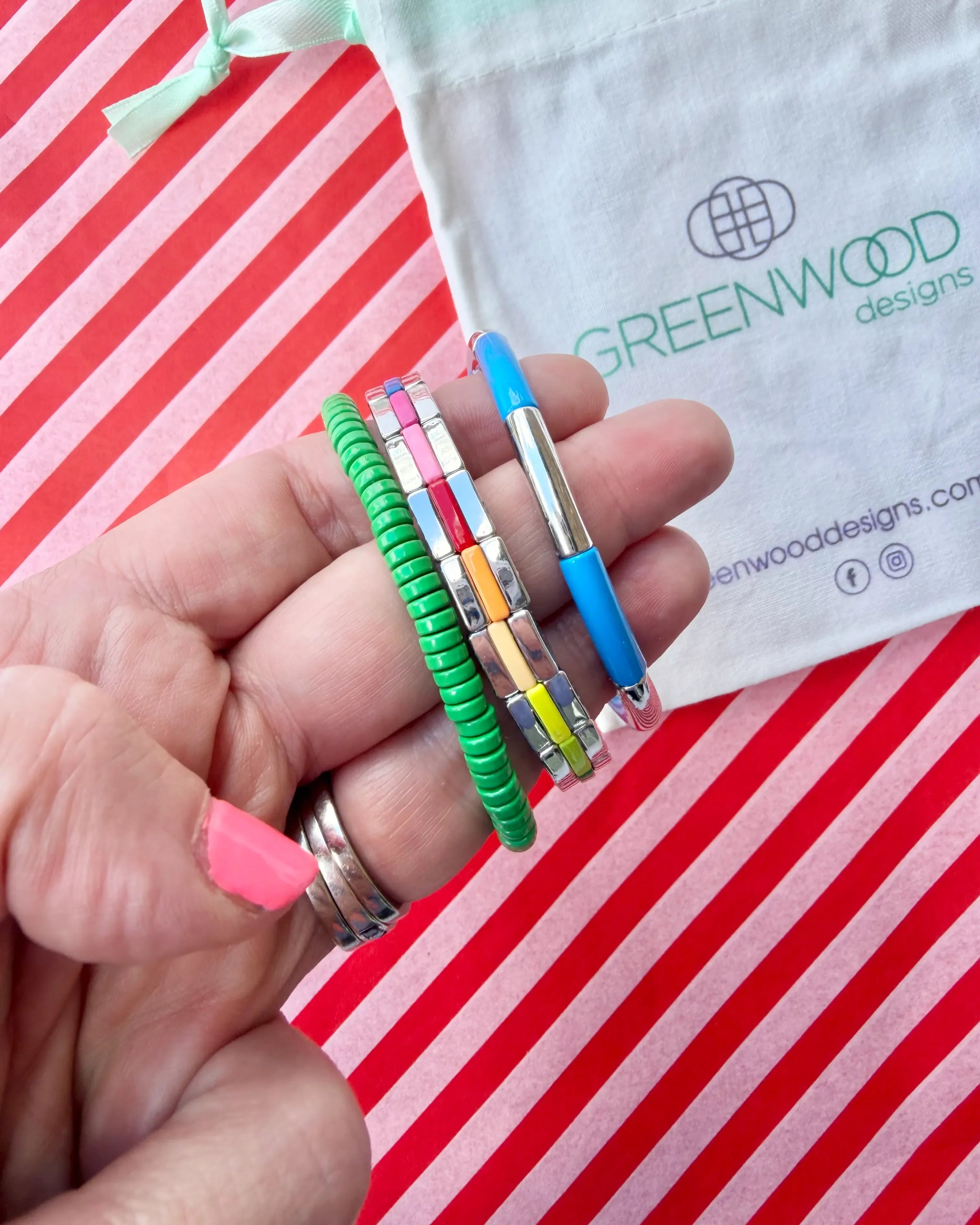 Hand holding three stacked bracelets: a green beaded bracelet, a silver tile bracelet with rainbow enamel stripes and a blue enamel tube bracelet with a silver bar, with a Greenwood Designs calico gift bag.