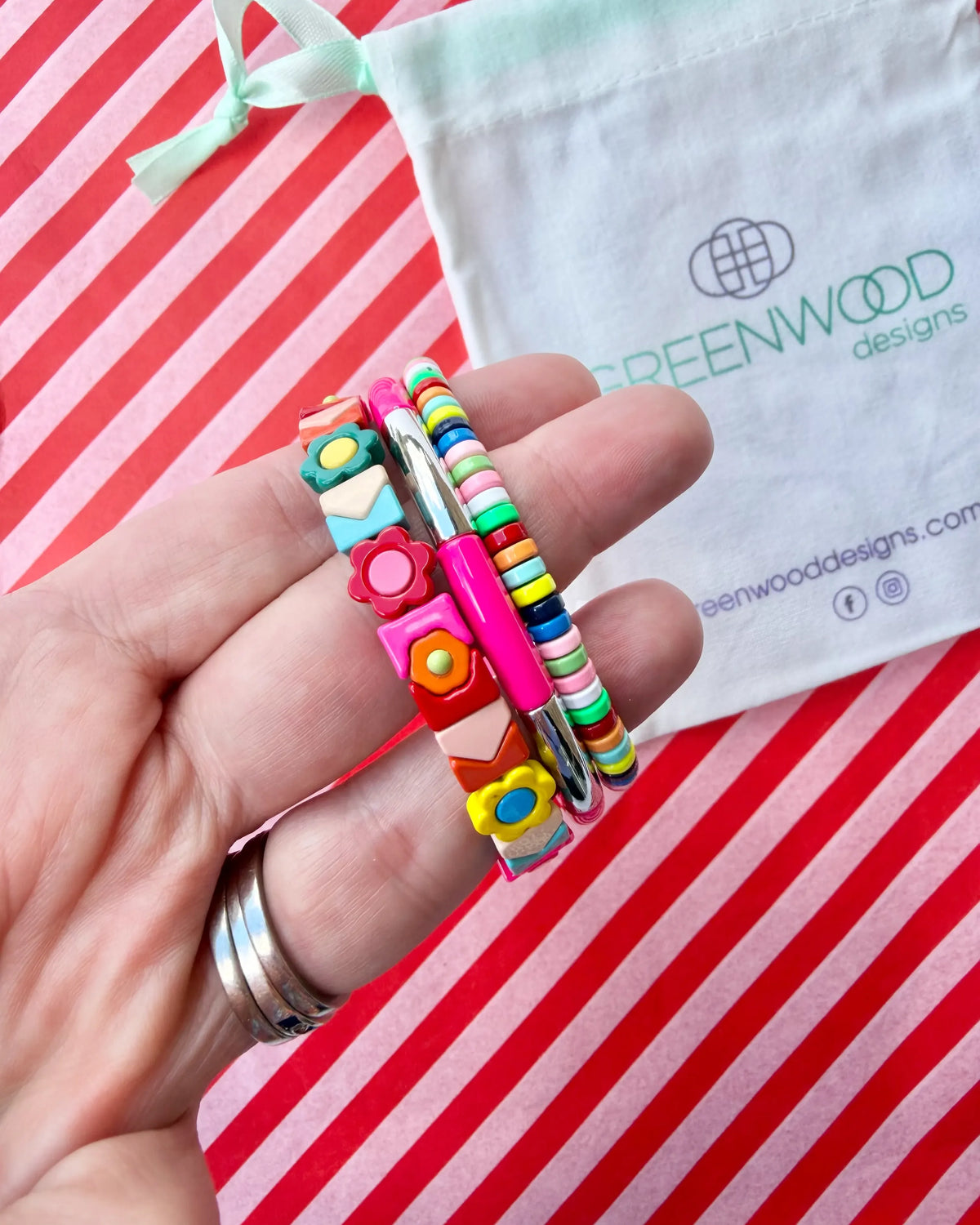 Hand holding three stacked bracelets: a multicoloured tile bracelet with flowers and hearts, a hot pink enamel tube bracelet with a silver bar and a rainbow beaded bracelet, with a Greenwood Designs calico gift bag.