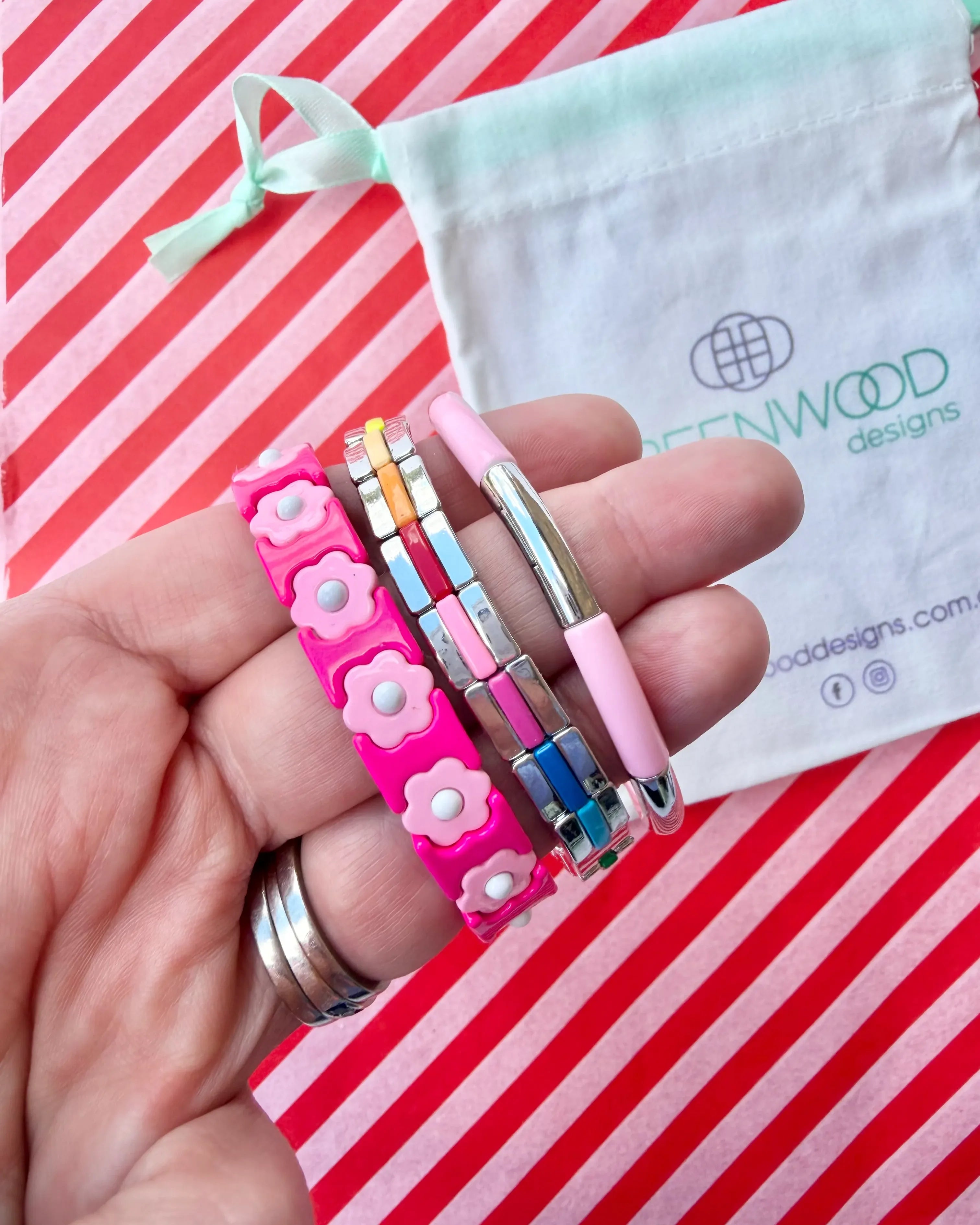 Hand holding three stacked bracelets: a hot pink enamel bracelet with pale pink flowers, a silver tile bracelet with rainbow enamel stripes and a pale pink enamel tube bracelet, with a Greenwood Designs calico gift bag.