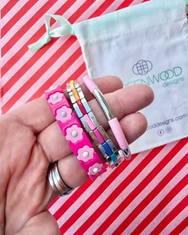 Hand holding three stacked bracelets: a hot pink enamel bracelet with pale pink flowers, a silver tile bracelet with rainbow enamel stripes and a pale pink enamel tube bracelet, with a Greenwood Designs calico gift bag.