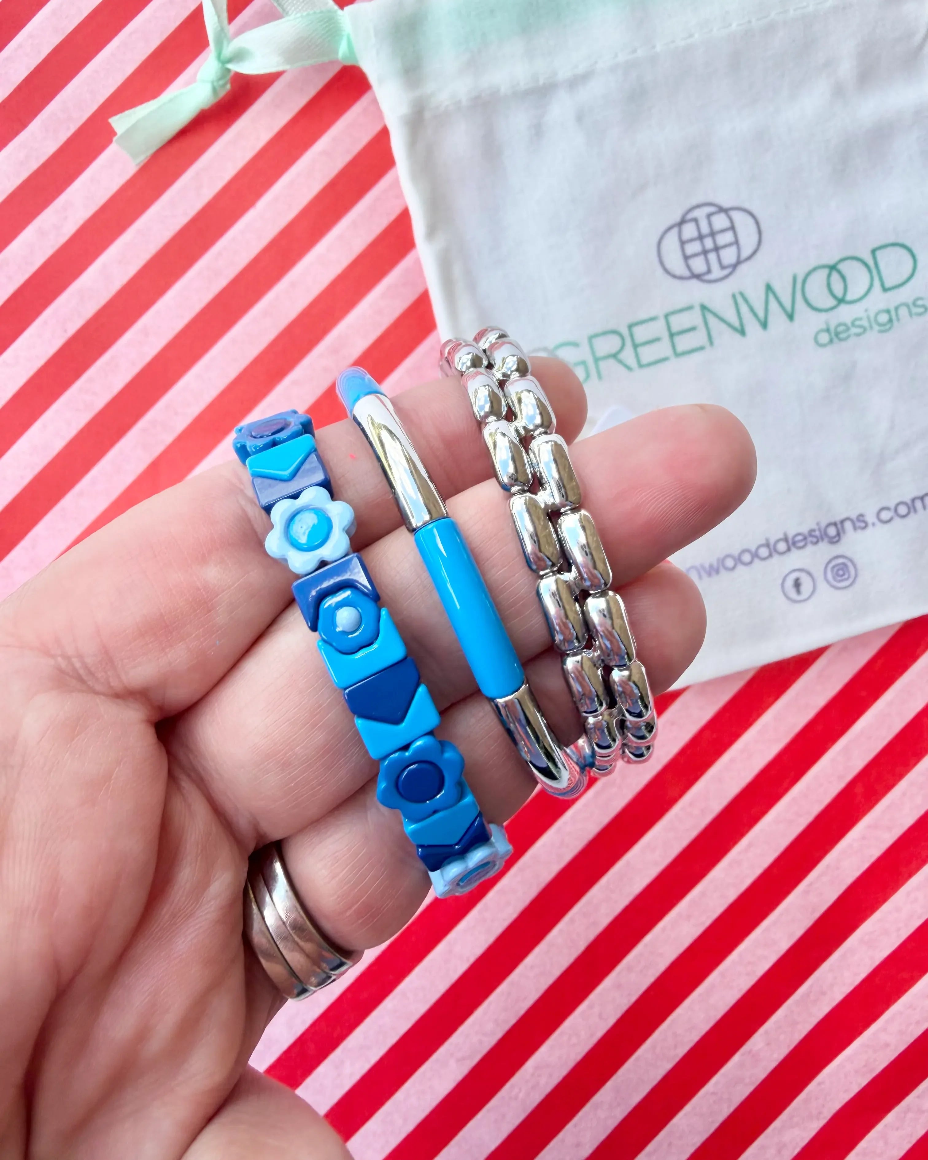 Hand holding three stacked bracelets: a blue and navy tile bracelet with flowers and hearts, a blue enamel tube bracelet with a silver bar and a chunky silver link bracelet, with a Greenwood Designs calico gift bag.
