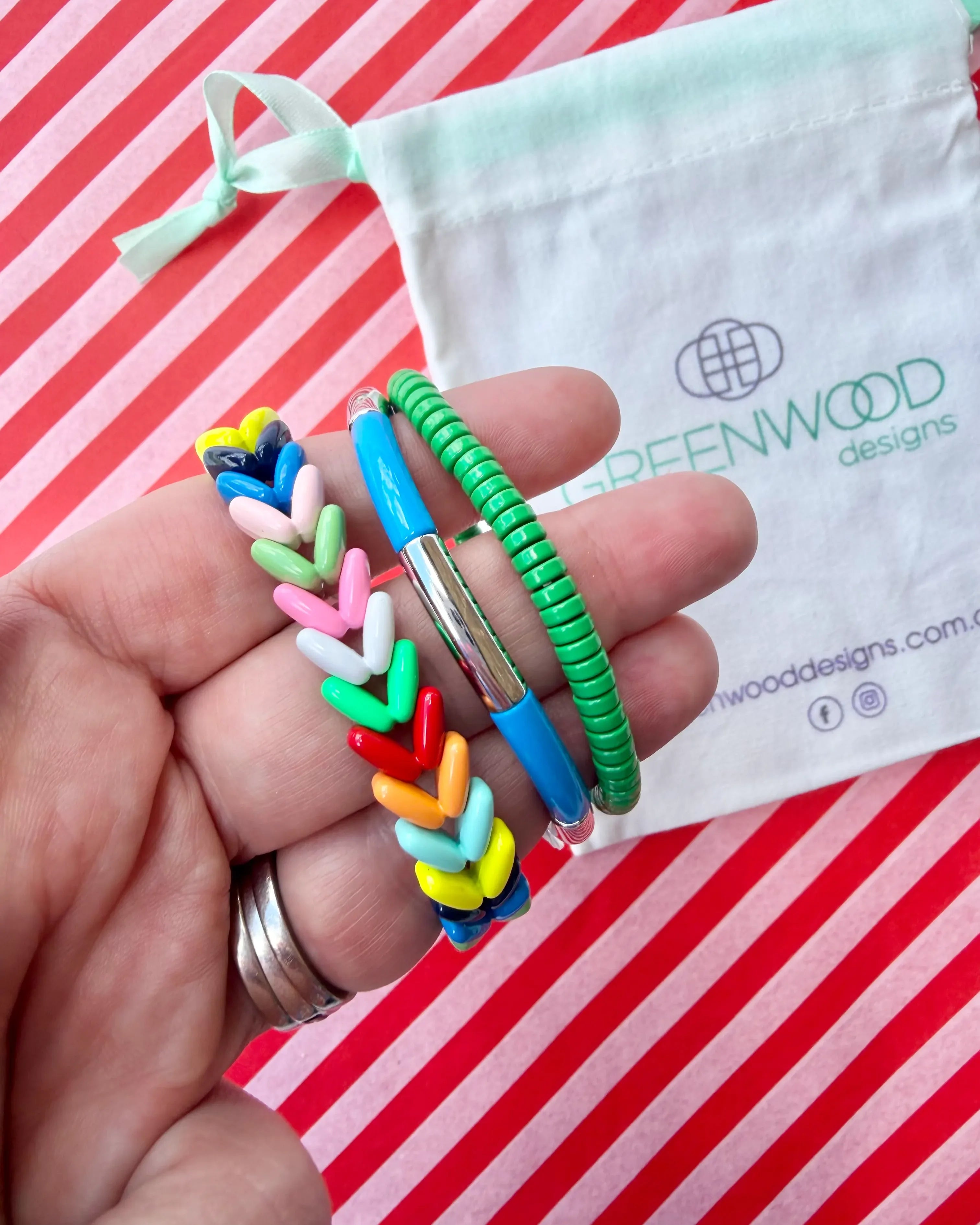 Hand holding three stacked bracelets: a rainbow multicoloured looped enamel bracelet, a blue enamel tube bracelet with a silver bar and a green beaded bracelet, with a Greenwood Designs calico gift bag.