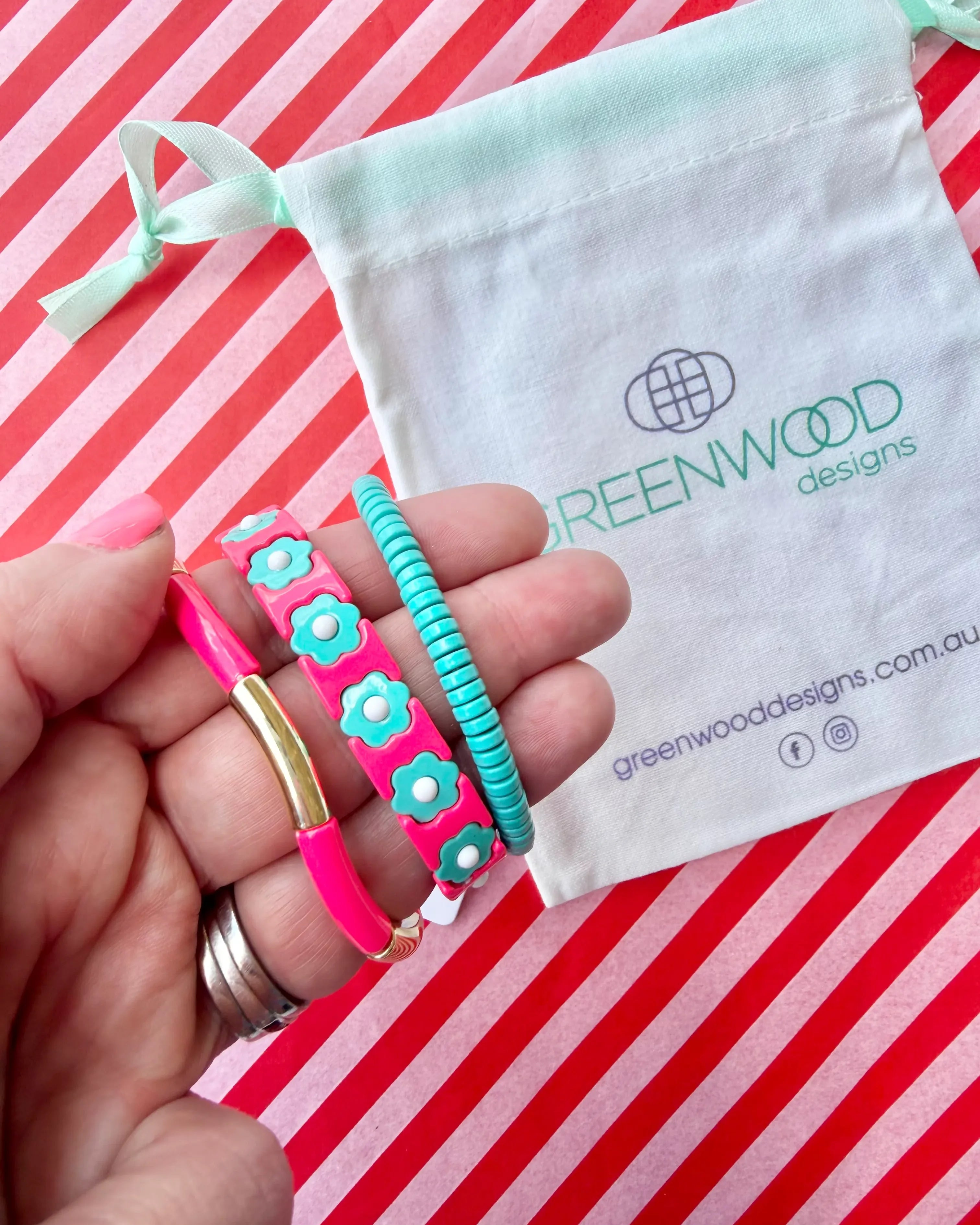 Hand holding three stacked bracelets: a coral pink enamel tube bracelet with a gold bar, a coral pink enamel bracelet with aqua flowers and an aqua beaded bracelet, with a Greenwood Designs calico gift bag.