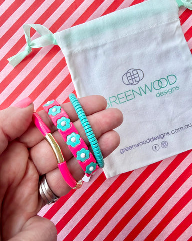 Hand holding three stacked bracelets: a coral pink enamel tube bracelet with a gold bar, a coral pink enamel bracelet with aqua flowers and an aqua beaded bracelet, with a Greenwood Designs calico gift bag.