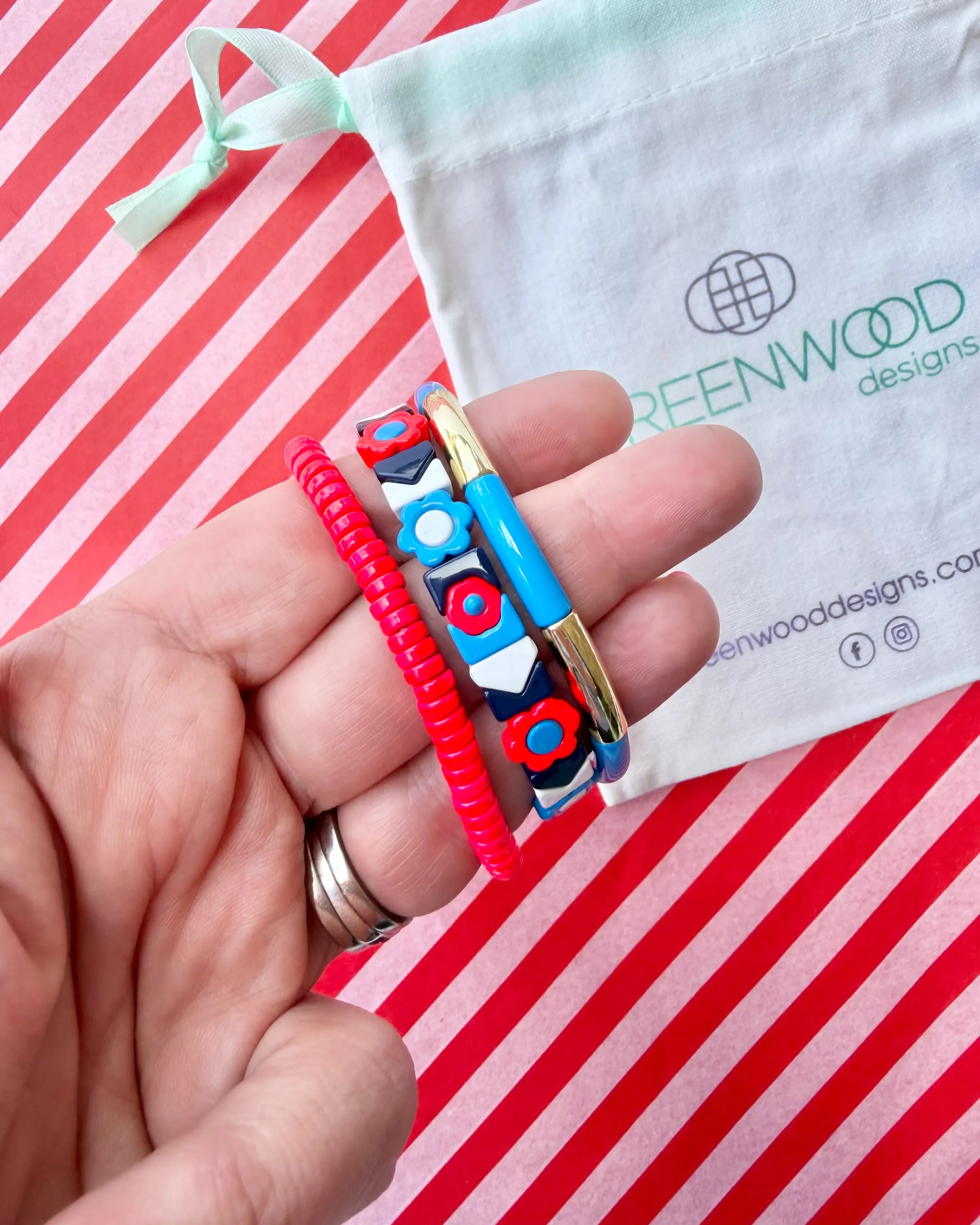 Hand holding three stacked bracelets: a red beaded bracelet, a navy tile bracelet with red and blue flowers and white chevrons, and a blue enamel tube bracelet with a gold bar, with a Greenwood Designs calico gift bag.