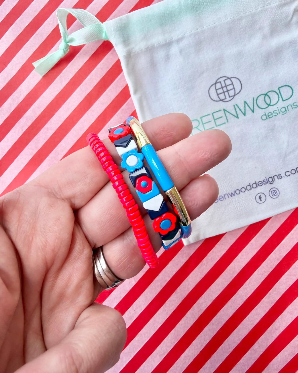 Hand holding three stacked bracelets: a red beaded bracelet, a navy tile bracelet with red and blue flowers and white chevrons, and a blue enamel tube bracelet with a gold bar, with a Greenwood Designs calico gift bag.