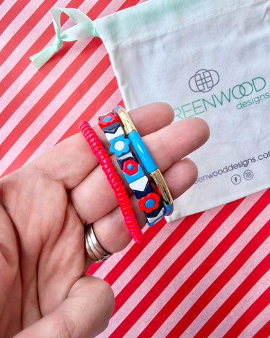 Hand holding three stacked bracelets: a red beaded bracelet, a navy tile bracelet with red and blue flowers and white chevrons, and a blue enamel tube bracelet with a gold bar, with a Greenwood Designs calico gift bag.