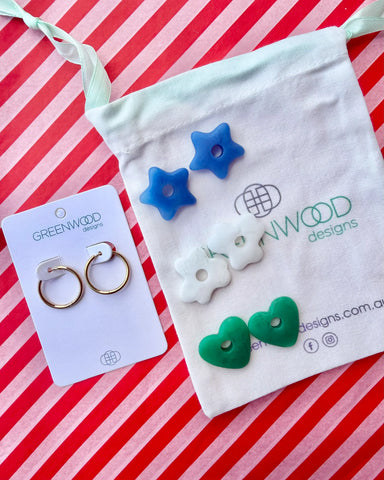 Hoop earrings on a Greenwood Designs card beside a calico pouch topped with three pairs of interchangeable resin charms: blue stars, white flowers and green hearts.
