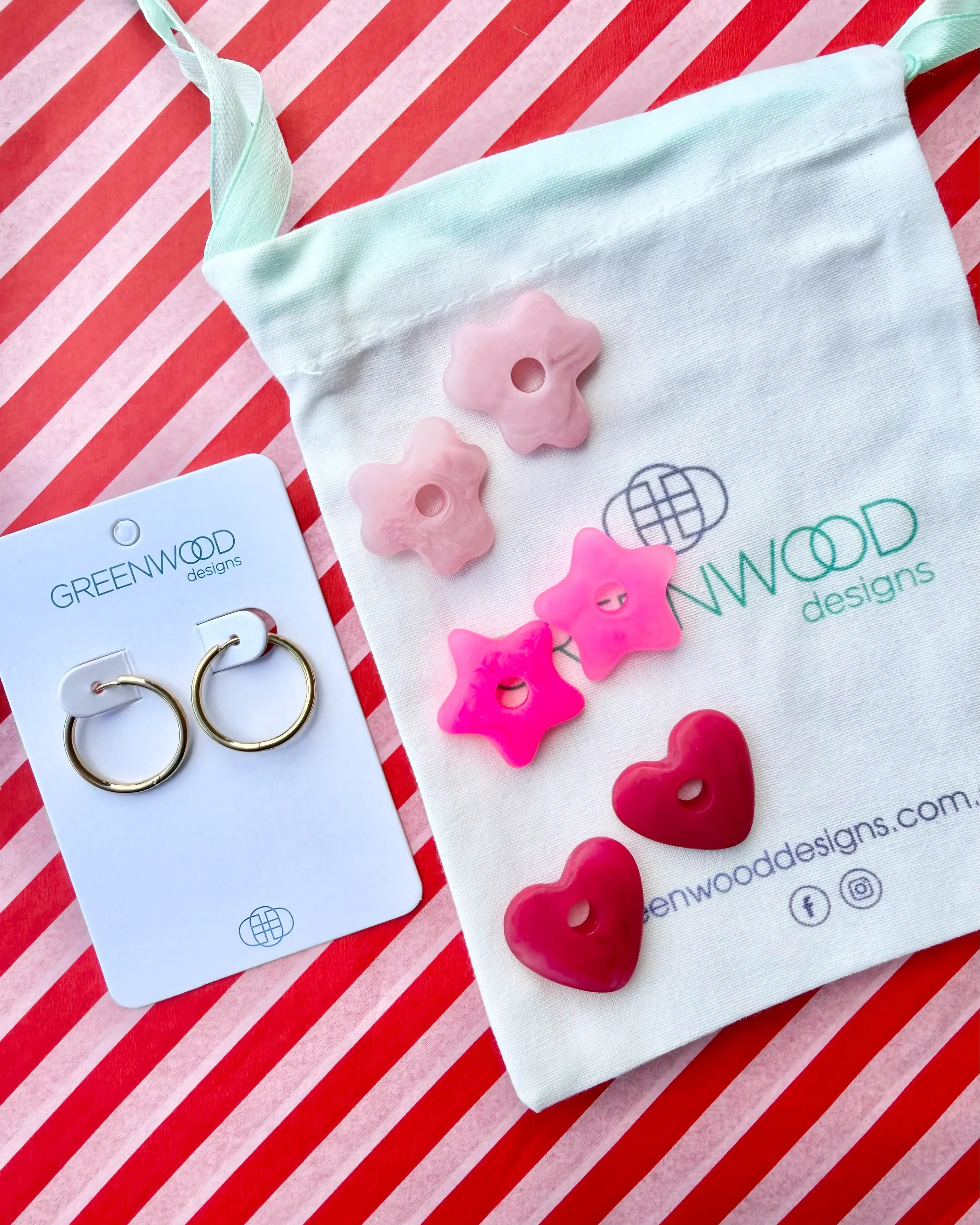 Hoop earrings on a Greenwood Designs card beside a calico pouch topped with three pairs of interchangeable resin charms: pale pink flowers, hot pink stars and raspberry red hearts.
