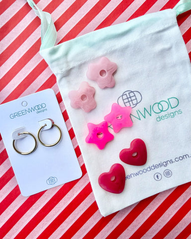 Hoop earrings on a Greenwood Designs card beside a calico pouch topped with three pairs of interchangeable resin charms: pale pink flowers, hot pink stars and raspberry red hearts.