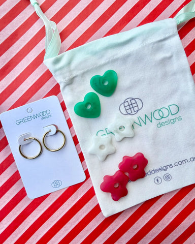 Hoop earrings on a Greenwood Designs card beside a calico pouch topped with three pairs of interchangeable resin charms: green hearts, white stars and raspberry red flowers.