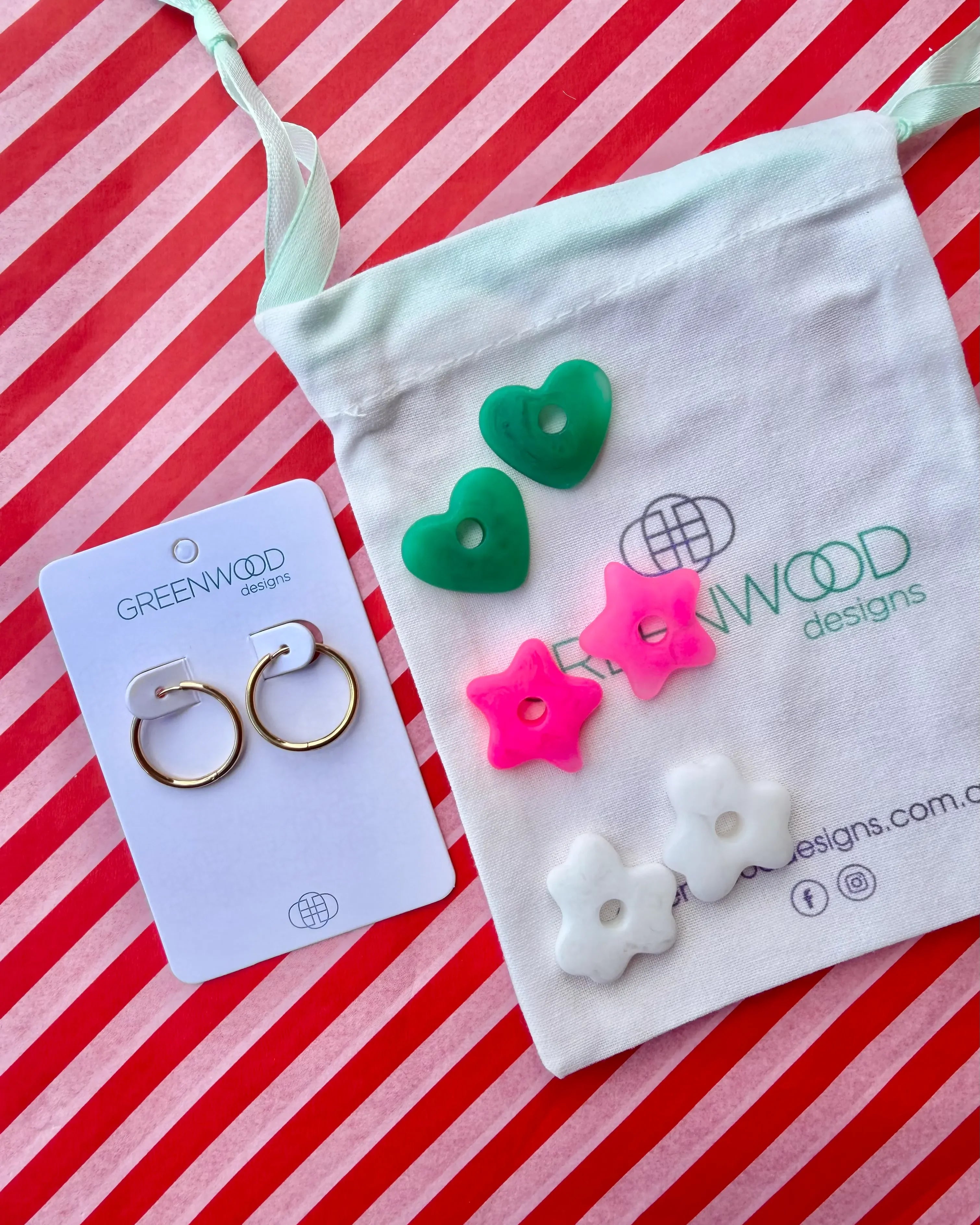 Hoop earrings on a Greenwood Designs card beside a calico pouch topped with three pairs of interchangeable resin charms: green hearts, hot pink stars and white flowers.