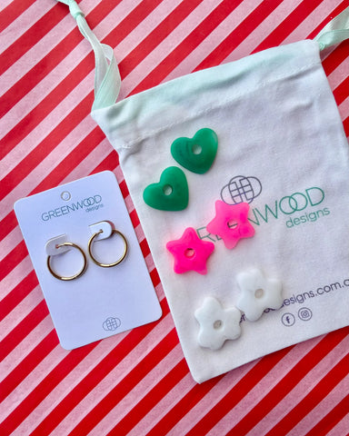 Hoop earrings on a Greenwood Designs card beside a calico pouch topped with three pairs of interchangeable resin charms: green hearts, hot pink stars and white flowers.