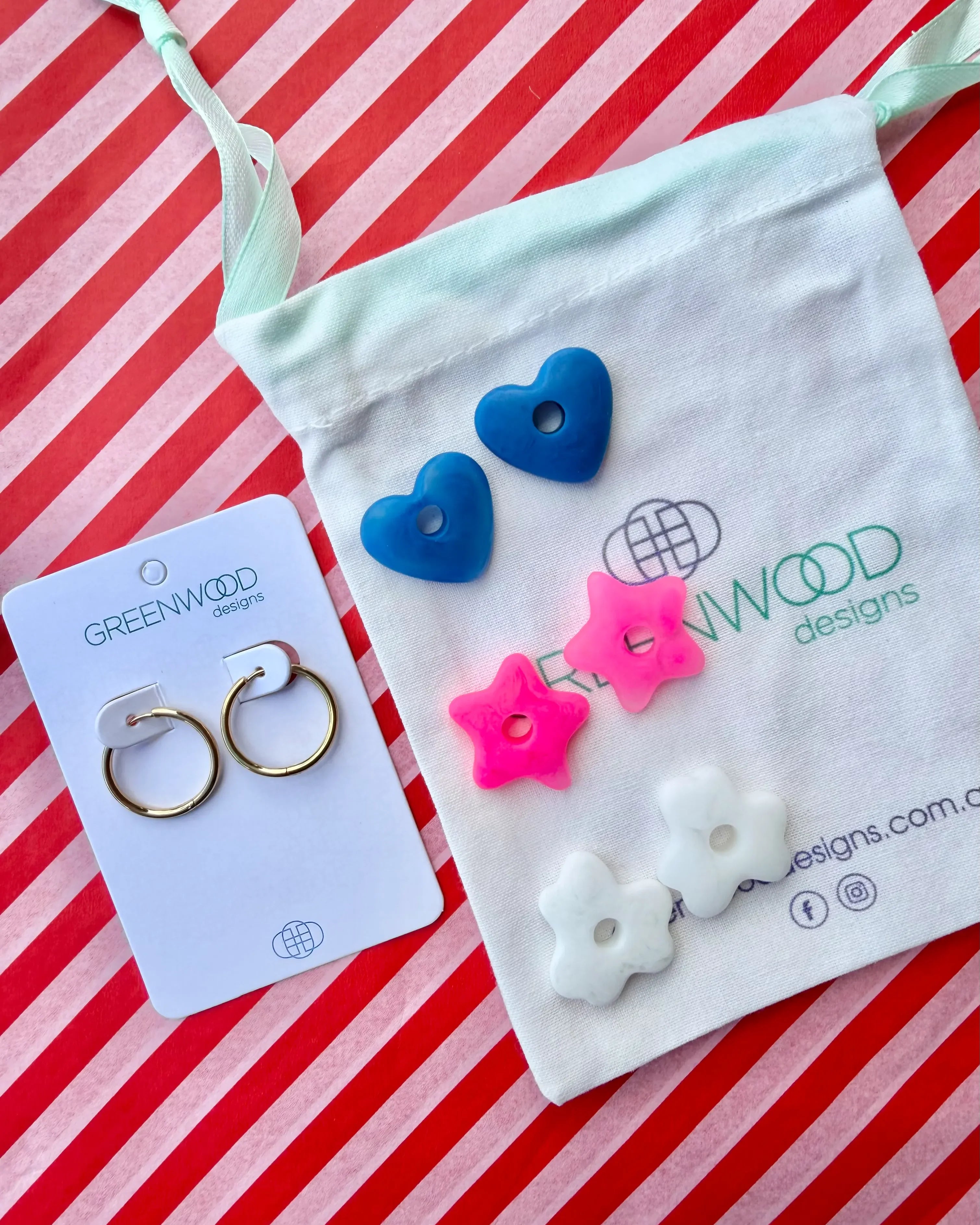 Hoop earrings on a Greenwood Designs card beside a calico pouch topped with three pairs of interchangeable resin charms: blue hearts, hot pink stars and white flowers.