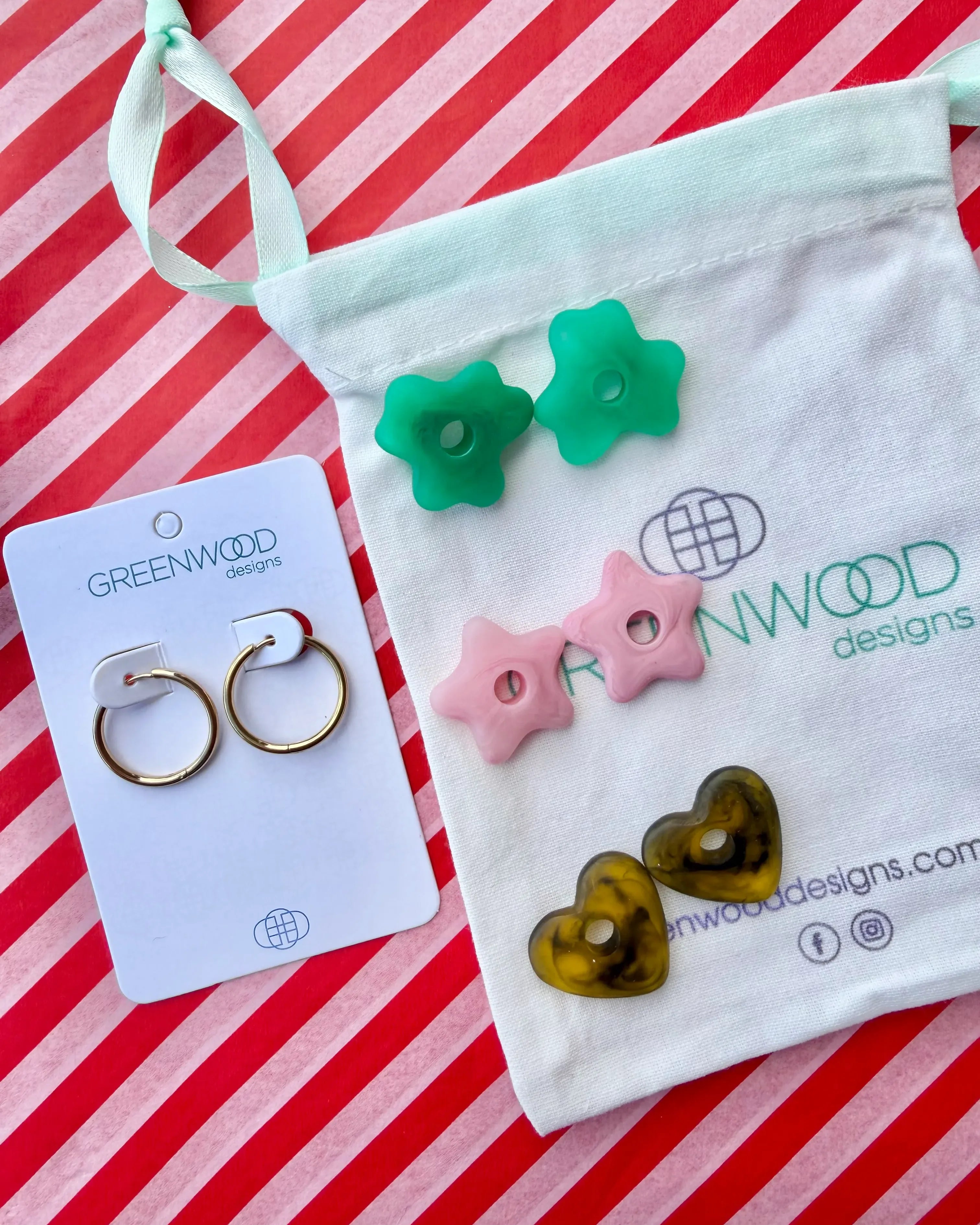 Hoop earrings on a Greenwood Designs card beside a calico pouch topped with three pairs of interchangeable resin charms: green flowers, pale pink stars and tortoiseshell hearts.