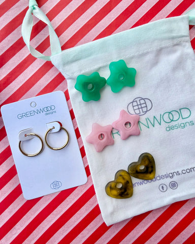 Hoop earrings on a Greenwood Designs card beside a calico pouch topped with three pairs of interchangeable resin charms: green flowers, pale pink stars and tortoiseshell hearts.