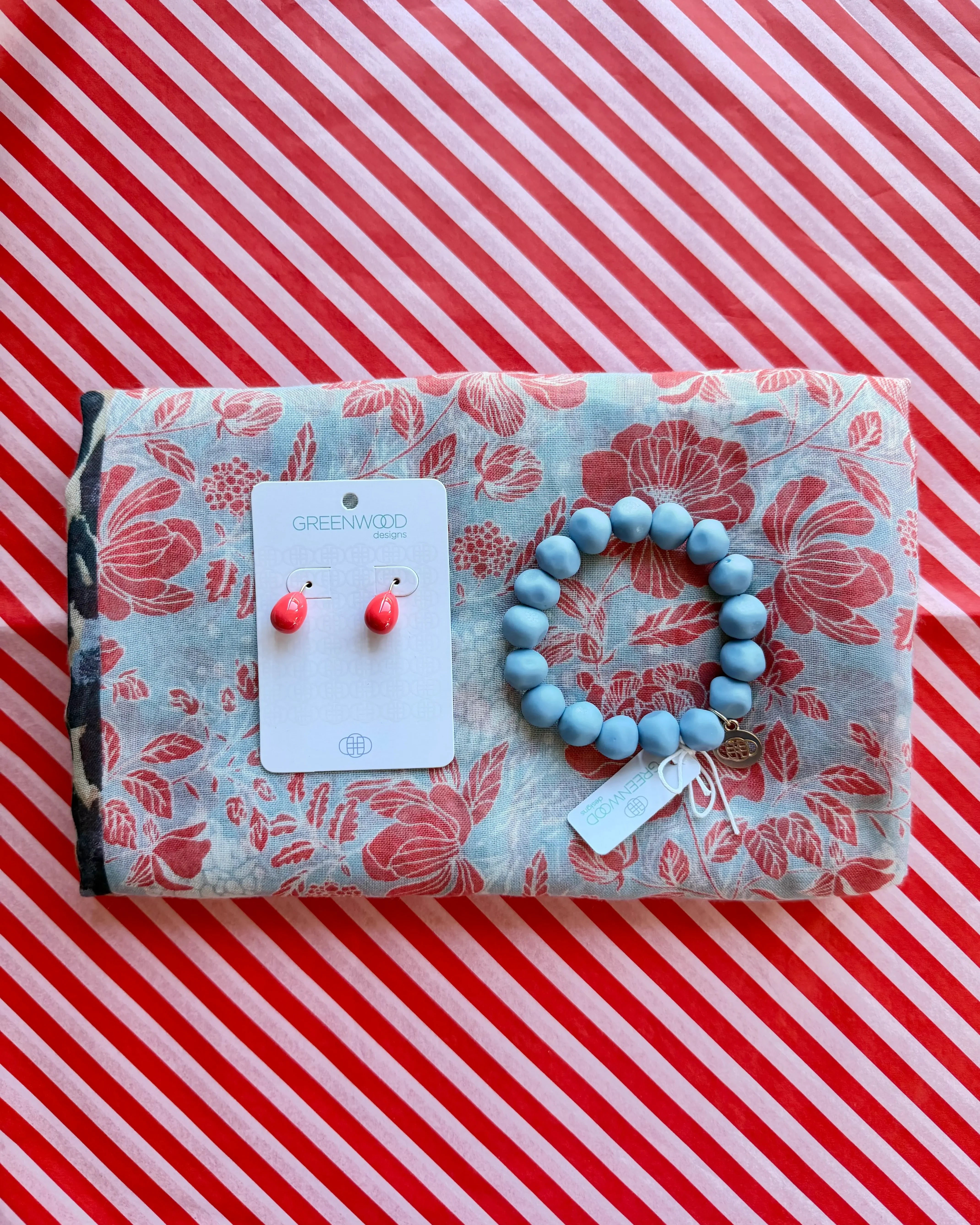 Flat lay of the Gemma scarf in pale blue with a coral red floral print, the Jacaranda rock bracelet and Alice earrings in Bright Coral on a Greenwood Designs card.