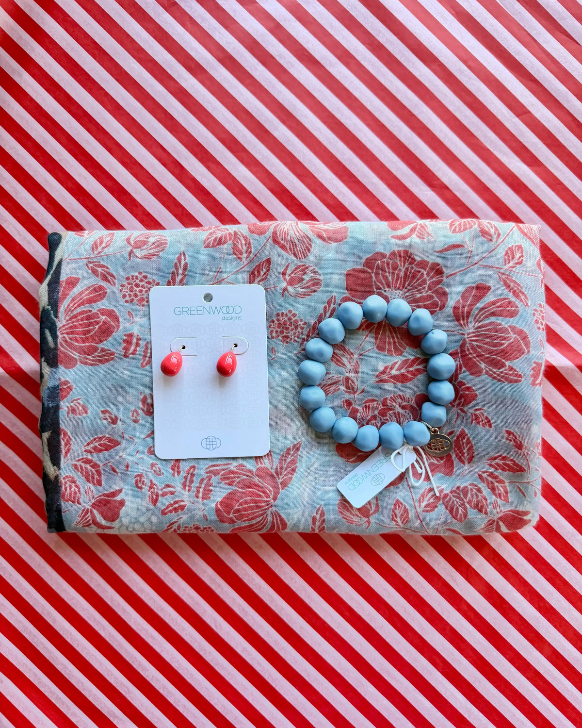 Flat lay of the Gemma scarf in pale blue with a coral red floral print, the Jacaranda rock bracelet and Alice earrings in Bright Coral on a Greenwood Designs card.