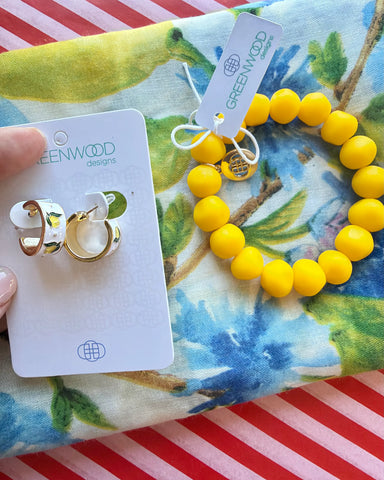 Hand holding Hayley earrings, gold and white enamel huggie hoops with a lemon print, on a Greenwood Designs card beside the Yellow rock bracelet, on the Iris scarf.