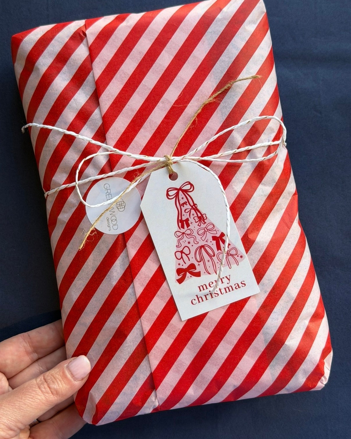 A Greenwood Designs gift wrapped in red and white striped tissue paper, tied with twine and a Merry Christmas gift tag with a bow-covered tree illustration.