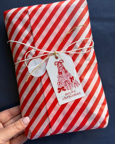 A Greenwood Designs gift wrapped in red and white striped tissue paper, tied with twine and a Merry Christmas gift tag with a bow-covered tree illustration.