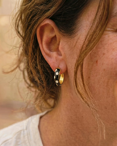 Woman wearing Jane black enamel chunky hoop earrings with white flower detail by Greenwood Designs, styled with a white top outdoors
