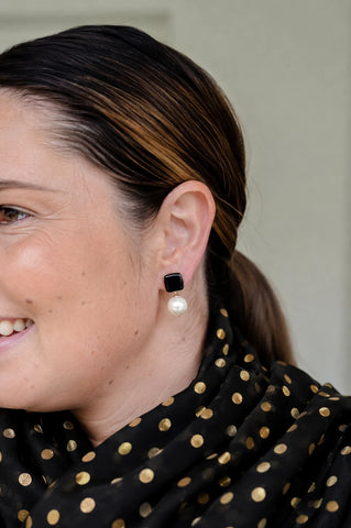 Woman smiling wearing Felicity black enamel square stud earrings with faux pearl drop by Greenwood Designs, styled with a black and gold top