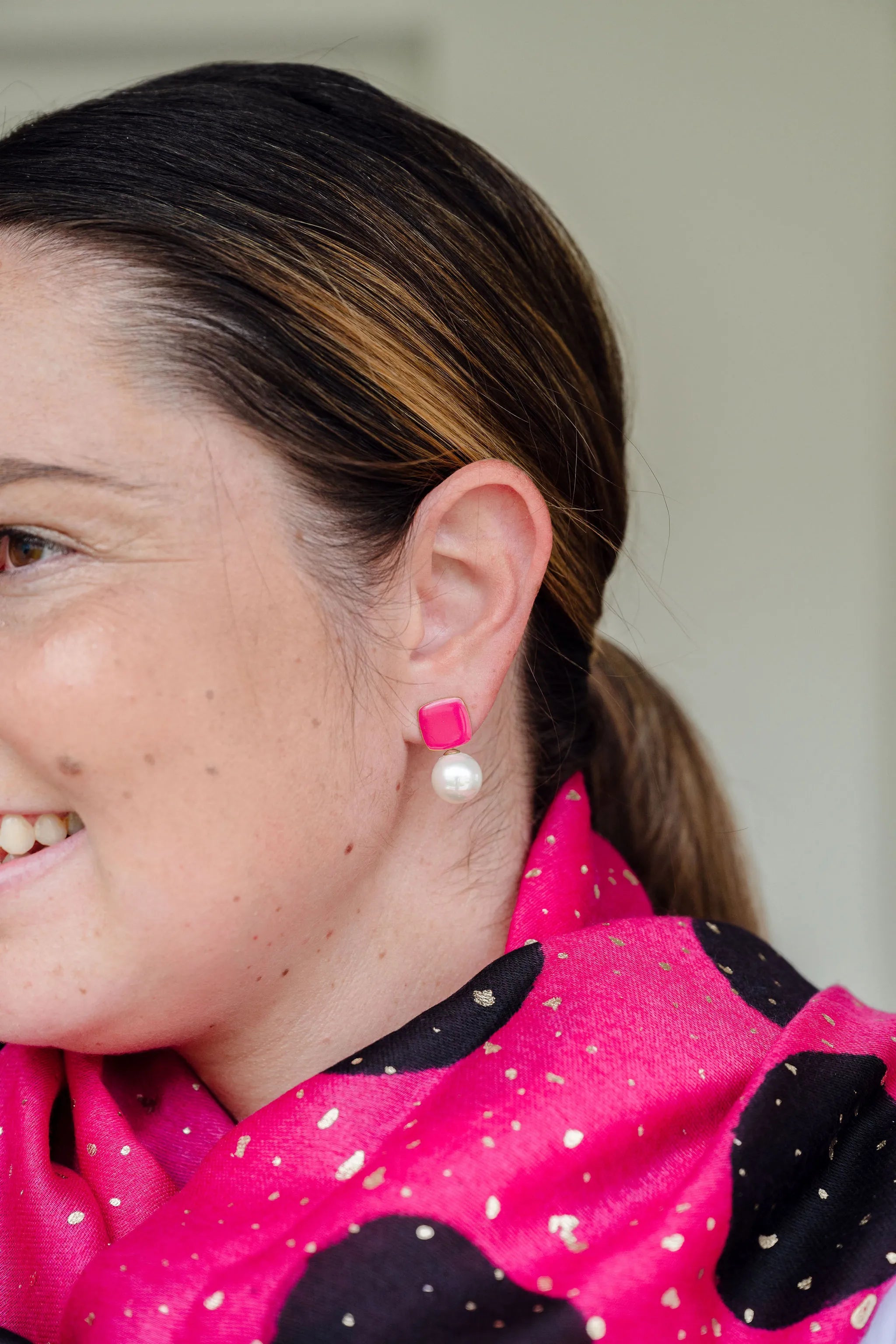 Woman smiling wearing Felicity bright pink enamel square stud earrings with faux pearl drop by Greenwood Designs, styled with a hot pink and black scarf