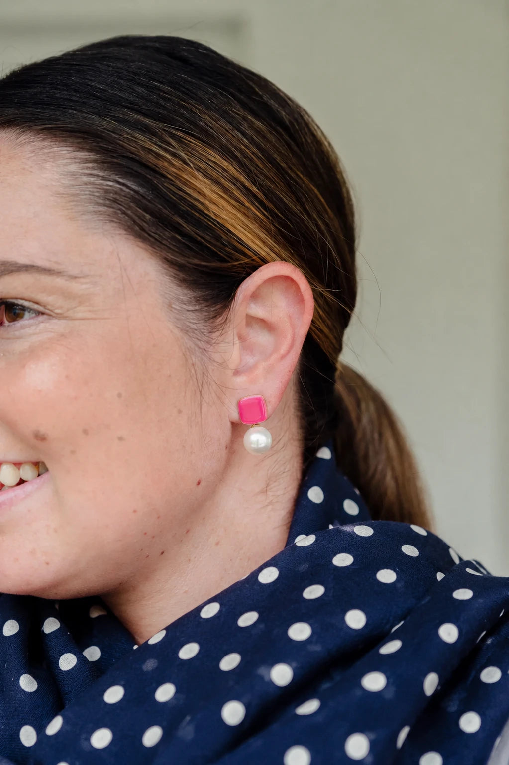 Woman smiling wearing Felicity light pink enamel square stud earrings with faux pearl drop by Greenwood Designs, styled with a navy blue polka dot top
