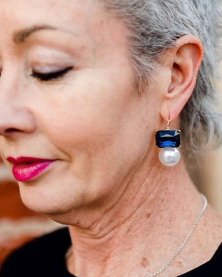 Older woman wearing navy blue rectangular crystal and faux pearl drop earrings by Greenwood Designs, styled with a black top