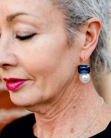 Older woman wearing navy blue rectangular crystal and faux pearl drop earrings by Greenwood Designs, styled with a black top