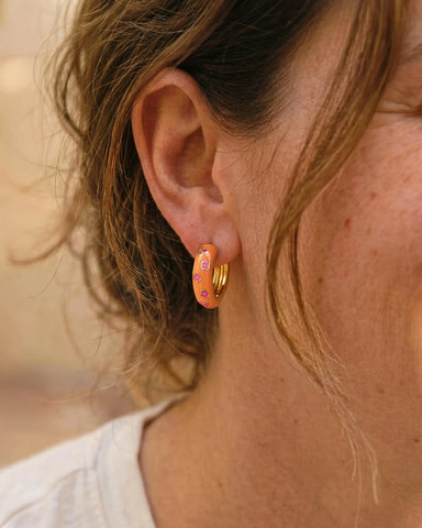 Woman wearing Jane orange enamel chunky hoop earrings with pink flower detail by Greenwood Designs, styled with a white top outdoors