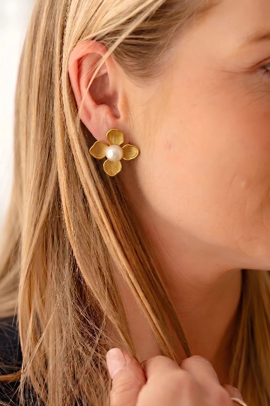 Close-up side profile of a woman with blonde hair wearing a gold four-petal flower stud earring featuring a large, round white pearl center. The gold petals have a brushed, textured finish with subtle vertical ridges.