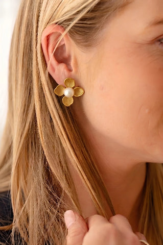 Close-up side profile of a woman with blonde hair wearing a gold four-petal flower stud earring featuring a large, round white pearl center. The gold petals have a brushed, textured finish with subtle vertical ridges.